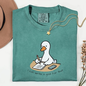 May include: Teal Comfort Colors t-shirt featuring a cartoon goose writing on a notepad with the text "I can write a goal for that." A gold star pendant necklace and a brown hat are also visible.