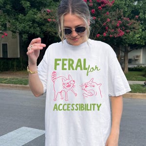 May include: White t-shirt with the text "FERAL for ACCESSIBILITY" in green and pink. The shirt features two cartoon cat illustrations in pink. The person in the image is wearing sunglasses and a gold bracelet.