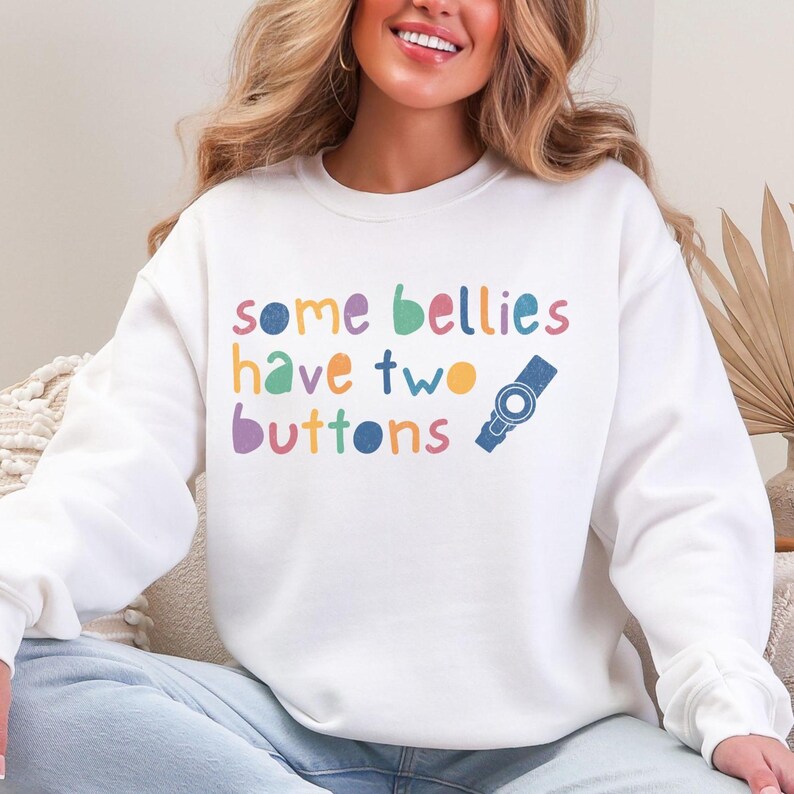 Puede incluir: Sudadera blanca con el texto "some bellies have two buttons" en colores arco&iacute;ris. Un bot&oacute;n azul se ilustra junto al texto.