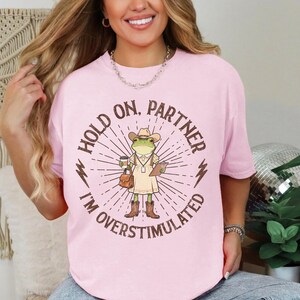 May include: Light pink t-shirt with a graphic of a frog wearing a cowboy hat, holding a coffee cup and clipboard. Text on the shirt reads "HOLD ON, PARTNER I'M OVERSTIMULATED" in a circular design with lightning bolt accents.