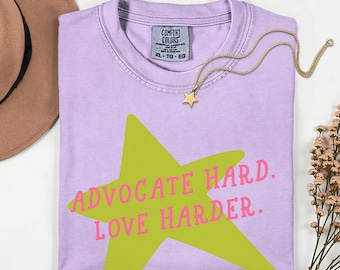 Può includere: Una t-shirt a maniche corte color viola chiaro con una grande grafica a stella verde lime e il testo rosa "ADVOCATE HARD. LOVE HARDER." stampato al centro. La maglietta presenta un girocollo e un'etichetta del marchio Comfort Colors.