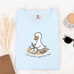 May include: Light blue t-shirt featuring a cartoon goose writing at a desk with the text "I can write a goal for that". The shirt has a crew neck and short sleeves. A coffee cup, sunglasses, and a notebook are also visible.