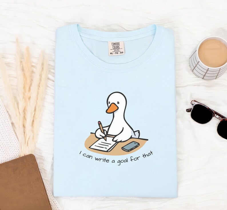 May include: Light blue t-shirt featuring a cartoon goose writing at a desk with the text "I can write a goal for that". The shirt has a crew neck and short sleeves. A coffee cup, sunglasses, and a notebook are also visible.