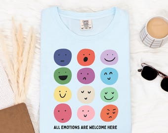 May include: Pale blue t-shirt featuring a variety of emoji faces in different colours, with the text "ALL EMOTIONS ARE WELCOME HERE." The shirt has a crew neck. A cup of coffee and sunglasses are in the background.