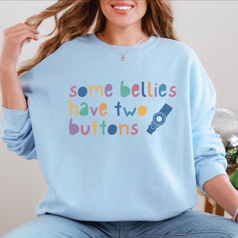 Puede incluir: Una sudadera azul claro con el texto "some bellies have two buttons" en colores arco&iacute;ris. Un bot&oacute;n azul con un c&iacute;rculo blanco est&aacute; al lado del texto.