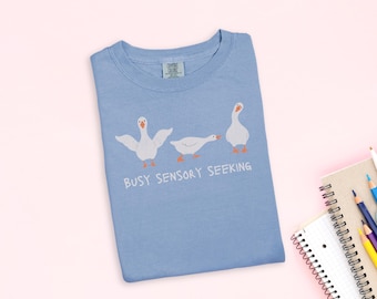 May include: A light blue t-shirt with a graphic of three white geese in different poses. The text "Busy Sensory Seeking" is printed below the geese. The shirt is folded on a pink surface.