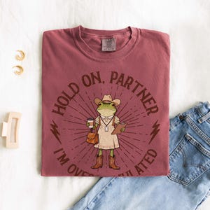 May include: A maroon t-shirt with the text "HOLD ON, PARTNER I'M OVERWHELMED" encircling a cartoon frog wearing a cowboy hat, dress, and boots. The frog is holding a coffee cup, a clipboard, and a purse. The shirt is paired with blue jeans.