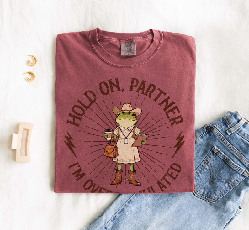May include: A maroon t-shirt with the text "HOLD ON, PARTNER I'M OVERWHELMED" encircling a cartoon frog wearing a cowboy hat, dress, and boots. The frog is holding a coffee cup, a clipboard, and a purse. The shirt is paired with blue jeans.