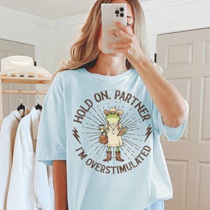 May include: Light blue t-shirt with the text "HOLD ON, PARTNER I'M OVERSTIMULATED" surrounding a cartoon frog wearing a cowboy hat, boots, and holding a coffee cup and clipboard. The shirt is a relaxed fit.