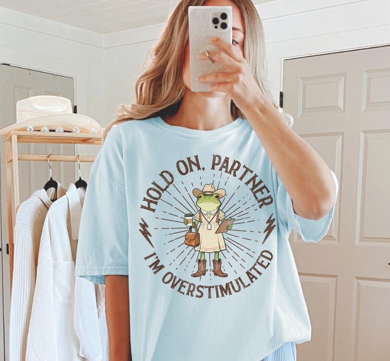 May include: Light blue t-shirt with the text "HOLD ON, PARTNER I'M OVERSTIMULATED" surrounding a cartoon frog wearing a cowboy hat, boots, and holding a coffee cup and clipboard. The shirt is a relaxed fit.