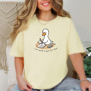 May include: A light yellow t-shirt featuring a cartoon goose writing on a notepad. The goose is sitting at a desk with a phone. The text on the shirt reads "I can write a goal for that."