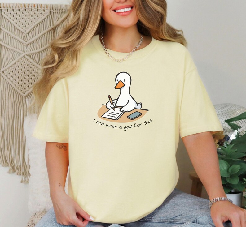 May include: A light yellow t-shirt featuring a cartoon goose writing on a notepad. The goose is sitting at a desk with a phone. The text on the shirt reads "I can write a goal for that."