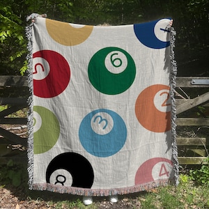 May include: A woven throw blanket with a white background and a pattern of colorful billiard balls with numbers. The balls are in various colors, including red, green, blue, yellow, and orange.
