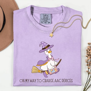 May include: A light purple t-shirt with a cartoon goose wearing a witch hat and cape, riding a broom. The goose holds a tablet and the text "ON MY WAY TO CHARGE AAC DEVICES" is printed at the bottom. A gold star necklace is draped across the shirt.