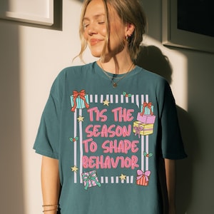 May include: Teal-colored t-shirt with the phrase "Tis the Season to Shape Behavior" in pink and white. The design includes illustrations of wrapped gifts, stars, and holly sprigs, creating a festive and playful look.