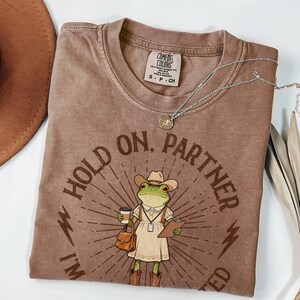 May include: Brown Comfort Colors t-shirt with a graphic of a frog wearing a cowboy hat, holding a coffee cup and clipboard. The text reads "Hold on, Partner, I'm Needed". A silver necklace and a brown hat are also visible.