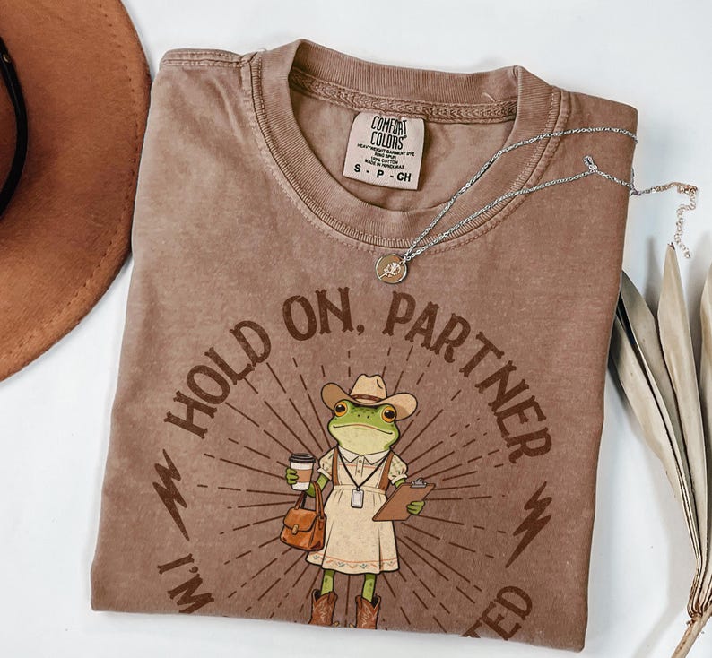 May include: Brown Comfort Colors t-shirt with a graphic of a frog wearing a cowboy hat, holding a coffee cup and clipboard. The text reads "Hold on, Partner, I'm Needed". A silver necklace and a brown hat are also visible.