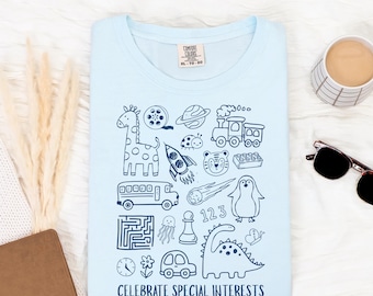 May include: Light blue t-shirt with a collection of whimsical, dark blue illustrations including a giraffe, rocket ship, and dinosaur. The text "CELEBRATE SPECIAL INTERESTS" is printed at the bottom. A coffee cup and sunglasses are in the background.
