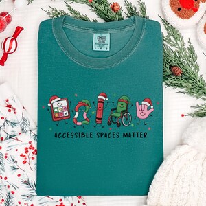 May include: A teal crewneck t-shirt featuring a graphic of five festive characters including a tablet, a candy cane, a crayon, a wheelchair, and a hand sign, all wearing Santa hats. The text below reads ACCESSIBLE SPACES MATTER in black.