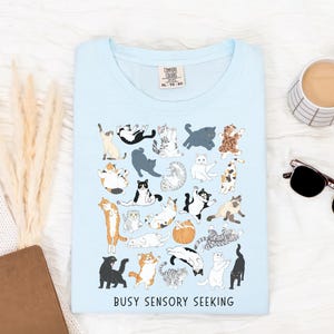 May include: Light blue t-shirt with a playful design of various cat illustrations in different poses. The shirt features the text "BUSY SENSORY SEEKING" at the bottom. The shirt is a Comfort Colors XL-TG-EG.
