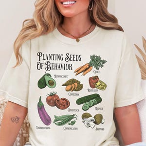 May include: Cream-coloured t-shirt featuring the text "Planting Seeds of Behaviour" and illustrations of vegetables. The design includes carrots, avocado, sweet potatoes, tomatoes, aubergine, peas, cucumbers, mushrooms, and bell peppers.