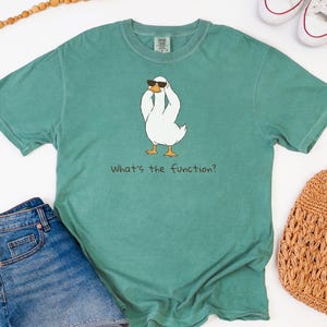 K&ouml;nnte beinhalten: Ein teal-farbenes T-Shirt mit einer Cartoon-Ente, die eine Sonnenbrille tr&auml;gt. Die wei&szlig;e Ente hat gelbe F&uuml;&szlig;e und h&auml;lt sich den Kopf. Der Text "What's the function?" steht darunter. Das Shirt wird mit Jeansshorts und einer gewebten Tasche kombiniert.