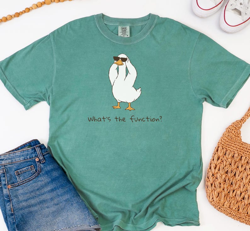 K&ouml;nnte beinhalten: Ein teal-farbenes T-Shirt mit einer Cartoon-Ente, die eine Sonnenbrille tr&auml;gt. Die wei&szlig;e Ente hat gelbe F&uuml;&szlig;e und h&auml;lt sich den Kopf. Der Text "What's the function?" steht darunter. Das Shirt wird mit Jeansshorts und einer gewebten Tasche kombiniert.