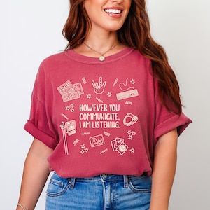 May include: A maroon t-shirt with the text "HOWEVER YOU COMMUNICATE, I AM LISTENING." The shirt features white illustrations of a keyboard, hand gestures, and other communication symbols. The shirt has short sleeves and a crew neck.