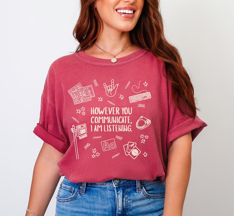 May include: A maroon t-shirt with the text "HOWEVER YOU COMMUNICATE, I AM LISTENING." The shirt features white illustrations of a keyboard, hand gestures, and other communication symbols. The shirt has short sleeves and a crew neck.