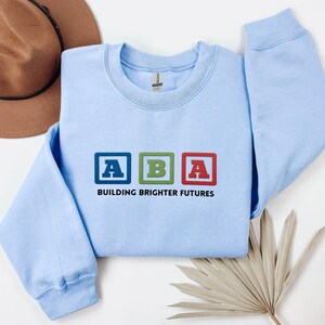 Puede incluir: Sudadera azul claro con las letras "ABA" en bloques de colores y el texto "BUILDING BRIGHTER FUTURES" debajo. La sudadera está hecha de un material suave.