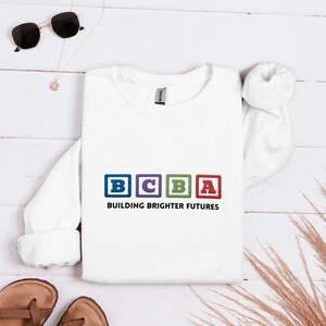 Puede incluir: Sudadera blanca con las letras "BCBA" en bloques coloridos, con el texto "BUILDING BRIGHTER FUTURES" debajo. La sudadera está colocada sobre una superficie de madera clara, con gafas de sol y sandalias cerca.