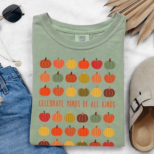 May include: Sage green t-shirt with a pumpkin graphic. The design features rows of colourful pumpkins and the text "CELEBRATE MINDS OF ALL KINDS". The shirt is folded and displayed with sunglasses, a necklace, jeans, and a sandal.