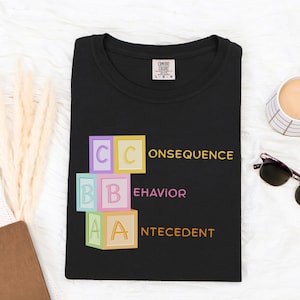 May include: Black t-shirt with the words "Consequence, Behavior, Antecedent" in yellow, pink, and orange. The letters are arranged as building blocks. A coffee cup and sunglasses are next to the shirt.