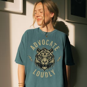 Peut inclure: T-shirt bleu sarcelle avec l'inscription "ADVOCATE LOUDLY" et une illustration de tigre. Le tigre est noir et beige, avec des éclairs de chaque côté. Le t-shirt a une coupe décontractée et est en matière douce.