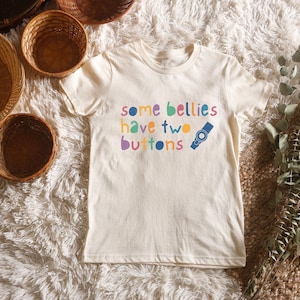 Peut inclure: T-shirt crème avec l'inscription colorée "some bellies have two buttons". Un dessin bleu et gris de montre est imprimé à côté du texte. Le t-shirt est présenté sur une surface blanche et moelleuse.