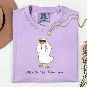 K&ouml;nnte beinhalten: Ein helllila Comfort Colors T-Shirt mit einer Cartoon-Ente, die eine Sonnenbrille tr&auml;gt und sich den Kopf h&auml;lt. Der Text "What's the function?" ist darunter gedruckt. Eine goldene Stern-Anh&auml;nger-Halskette liegt auf dem Shirt.