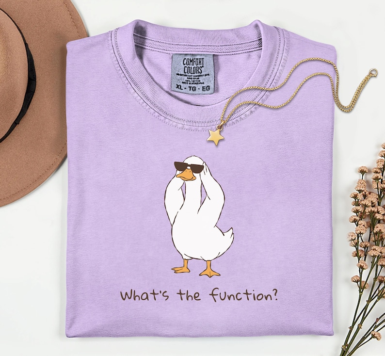 K&ouml;nnte beinhalten: Ein helllila Comfort Colors T-Shirt mit einer Cartoon-Ente, die eine Sonnenbrille tr&auml;gt und sich den Kopf h&auml;lt. Der Text "What's the function?" ist darunter gedruckt. Eine goldene Stern-Anh&auml;nger-Halskette liegt auf dem Shirt.