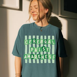 Peut inclure: Un t-shirt à manches courtes bleu sarcelle avec un motif à rayures verticales vert clair. Le texte CEREBRAL PALSY AWARENESS est imprimé en lettres capitales blanches sur des blocs rectangulaires vert foncé superposés au fond rayé.