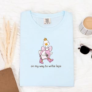 May include: Pale blue t-shirt with a cartoon goose wearing a crown, pink dress, and boots, holding a clipboard. The text "on my way to write ieps" is printed below the goose. The shirt has a crew neck.