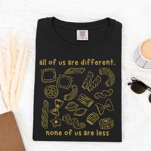 May include: Black t-shirt with a gold illustration of various pasta shapes and the text "all of us are different, none of us are less." The shirt has a crew neck and a small tag on the upper left.