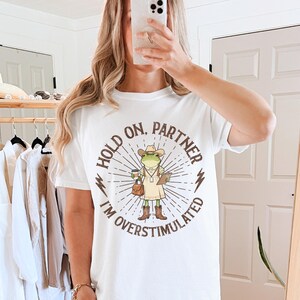 May include: White t-shirt with a graphic of a frog wearing a cowboy hat, holding a coffee cup and a clipboard. The text on the shirt reads "HOLD ON, PARTNER I'M OVERSTIMULATED" in a circular design with sunburst rays.