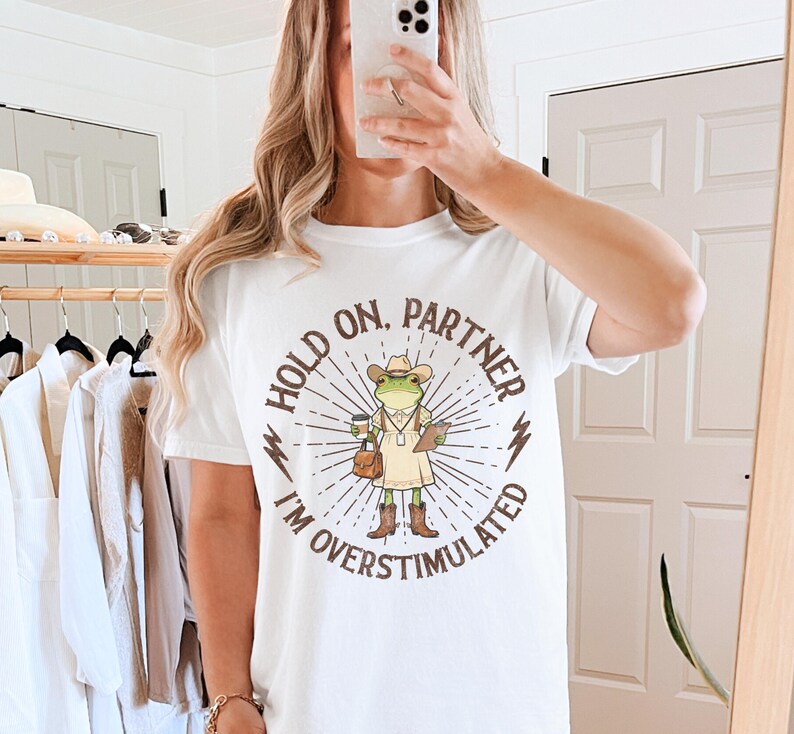 May include: White t-shirt with a graphic of a frog wearing a cowboy hat, holding a coffee cup and a clipboard. The text on the shirt reads "HOLD ON, PARTNER I'M OVERSTIMULATED" in a circular design with sunburst rays.