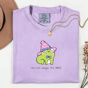 May include: A light purple t-shirt with a cartoon frog wearing a pink wizard hat and bow tie. The text "It's not magic. It's ABA." is printed below the frog. A gold necklace with a star pendant is draped across the shirt.