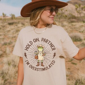 May include: Beige t-shirt with a graphic of a frog wearing a cowboy hat, holding a coffee cup and a notepad. The text on the shirt reads "Hold on, Partner, I'm Overstimulated". The shirt is a casual wear item.