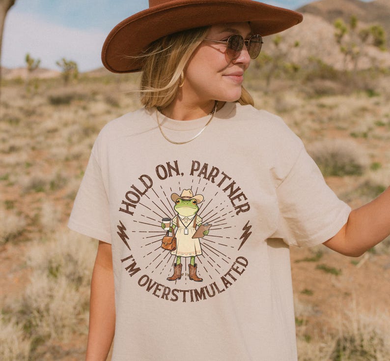 May include: Beige t-shirt with a graphic of a frog wearing a cowboy hat, holding a coffee cup and a notepad. The text on the shirt reads "Hold on, Partner, I'm Overstimulated". The shirt is a casual wear item.
