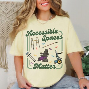 May include: A light yellow t-shirt with the text "Accessible Spaces Matter" in green and white. The shirt features illustrations of mobility aids, including a walker, wheelchair, and cane.