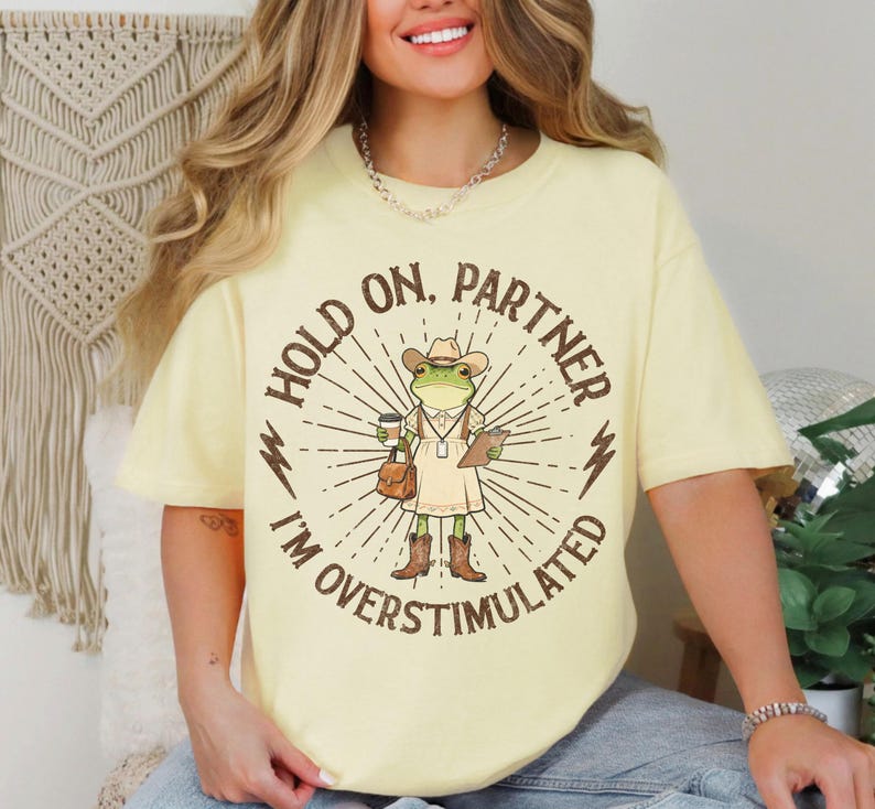 May include: A light yellow t-shirt with the text "HOLD ON, PARTNER I'M OVERSTIMULATED" surrounding a cartoon frog wearing a cowboy hat, dress, and boots. The frog is holding a coffee cup and a clipboard. The shirt has short sleeves.