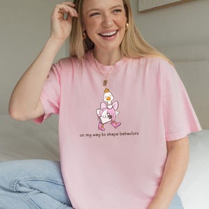 May include: A light pink t-shirt featuring a cartoon goose wearing a crown, dress, and boots. The goose is holding a briefcase and walking. The text "on my way to shape behaviors" is printed below the goose.