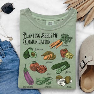 May include: Sage green t-shirt with the text "Planting Seeds of Communication" and illustrations of vegetables. The design includes an avocado, sweet potato, tomato, cucumber, and other produce.