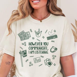 May include: Cream-colored t-shirt with the text "HOWEVER YOU COMMUNICATE, I AM LISTENING." and various green illustrations, including a keyboard, hand sign, and cards. The shirt has a relaxed fit.